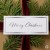Biała etykieta prezentowa ze świątecznym napisem Merry Christmas w kolorze srebrnym i ze srebrną ramka. Pod naklejką prezent z bordową wstażką i gałązką świerku. Prezent w stylu eko.