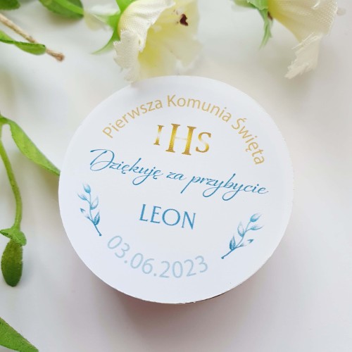 Okrągła personalizowana naklejka na Pierwszą Komunię Świętą dla chłopca. Widoczny złoty symbol IHS, niebieskie podziękowanie 'Dziękuję za przybycie', imię Leon oraz data 03.06.2023, ozdobiona niebieskimi gałązkami na jasnym tle.