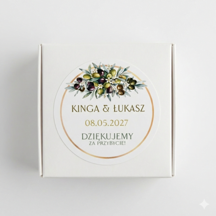 Białe kwadratowe pudełeczko na prezenty dla gości weselnych. Na wierzchu okrągła personalizowana naklejka ślubna w stylu toskańskim z motywem gałązek oliwnych, zielonymi oliwkami i złotą ramką, z imionami pary młodej oraz datą wesela.